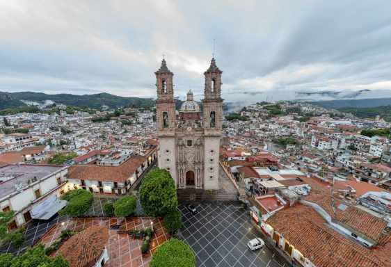 Taxco View of a cityscape featuring twin bell towers of a church against a cloudy sky.