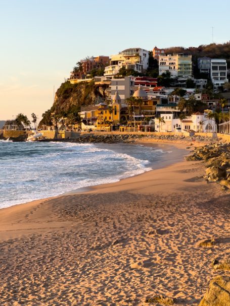 Mazatlan Scenic beach with gentle waves, rocky shore, and hillside houses at sunset.