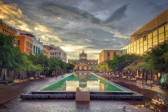 Guadalajara Historic plaza with a reflecting pool, surrounded by trees and buildings under a cloudy sky.