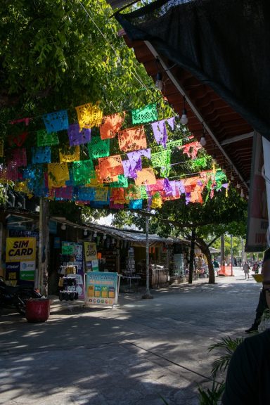Yaxchilan Colorful papel picado decorations hang over a quiet street with shops and trees.