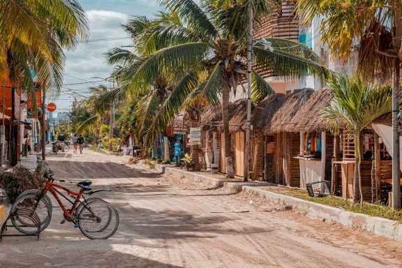 Isla Holbox Dirt road lined with palm trees and rustic huts, with a bicycle resting nearby.