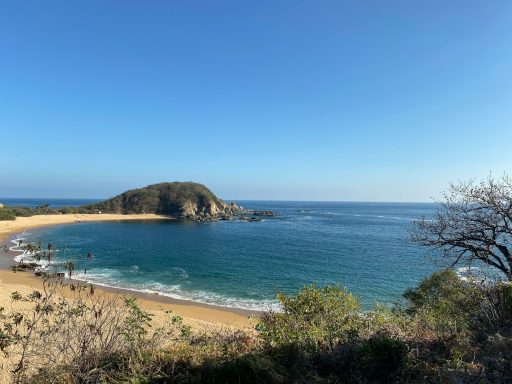 Huatulco Scenic beach view with calm water, sandy shore, and distant hills under a clear blue sky.
