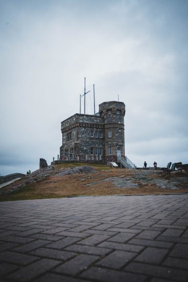 Stone tower with antennas, set against a cloudy sky and grassy terrain.